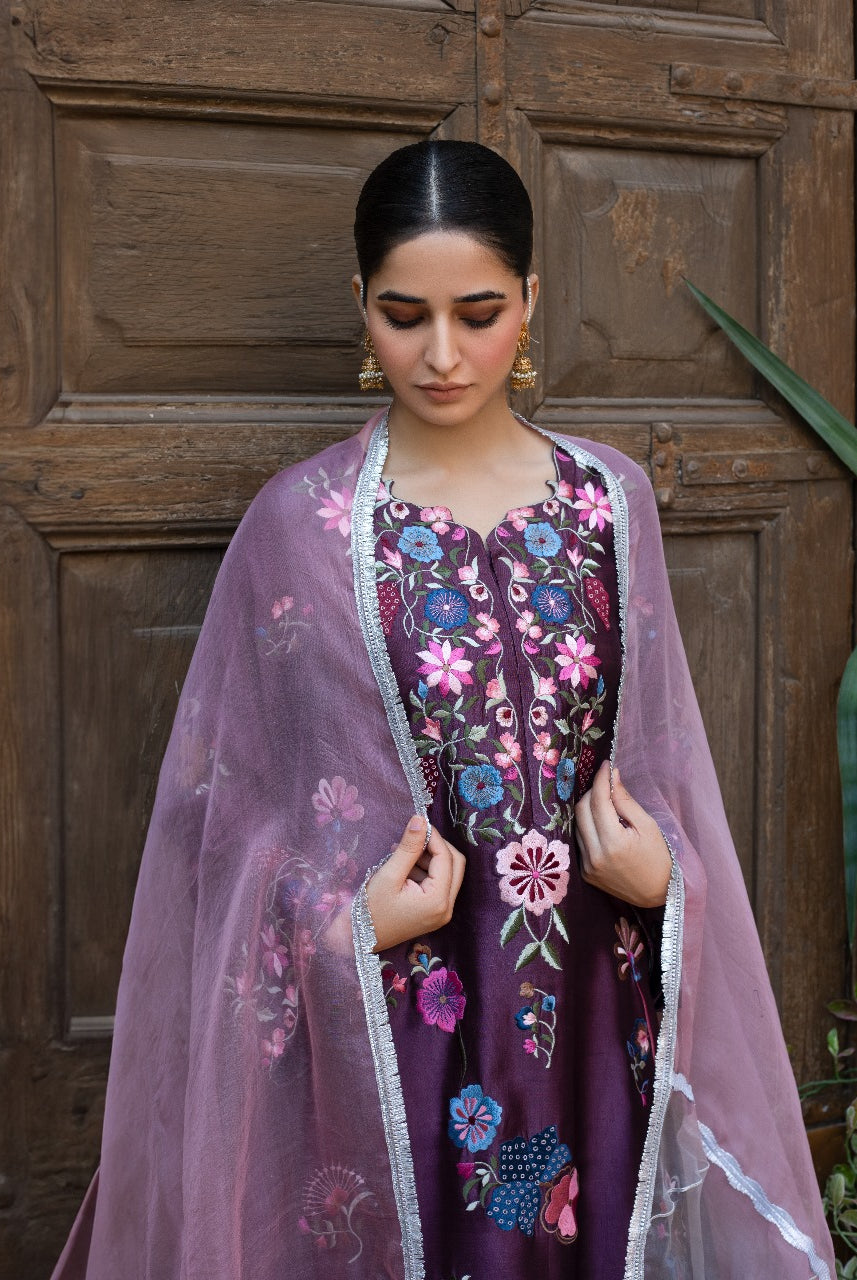 Woman wearing a traditional embroidered outfit with a pink dupatta in front of a wooden door.