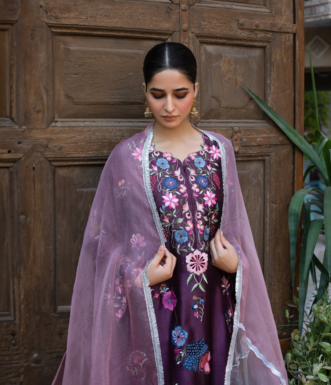 Woman wearing a traditional embroidered outfit with a pink dupatta in front of a wooden door.