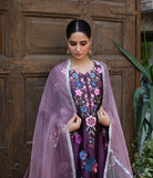 Woman wearing a traditional embroidered outfit with a pink dupatta in front of a wooden door.